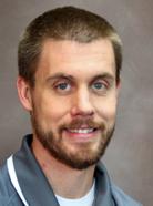 A man with short brown hair, a beard, and a mustache is wearing a gray collared shirt and smiling at the camera against a neutral brown background.